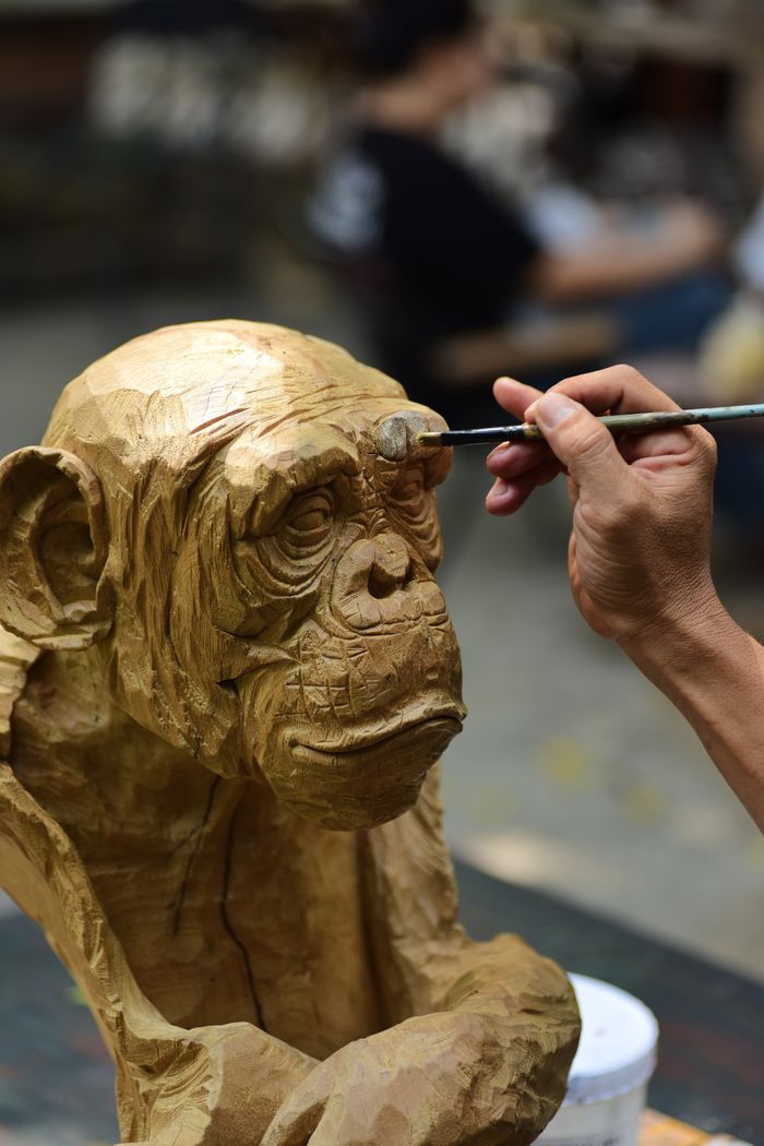social-feed-img-01 Close-up of an artist painting a wooden ape sculpture in a workshop, showcasing creativity.