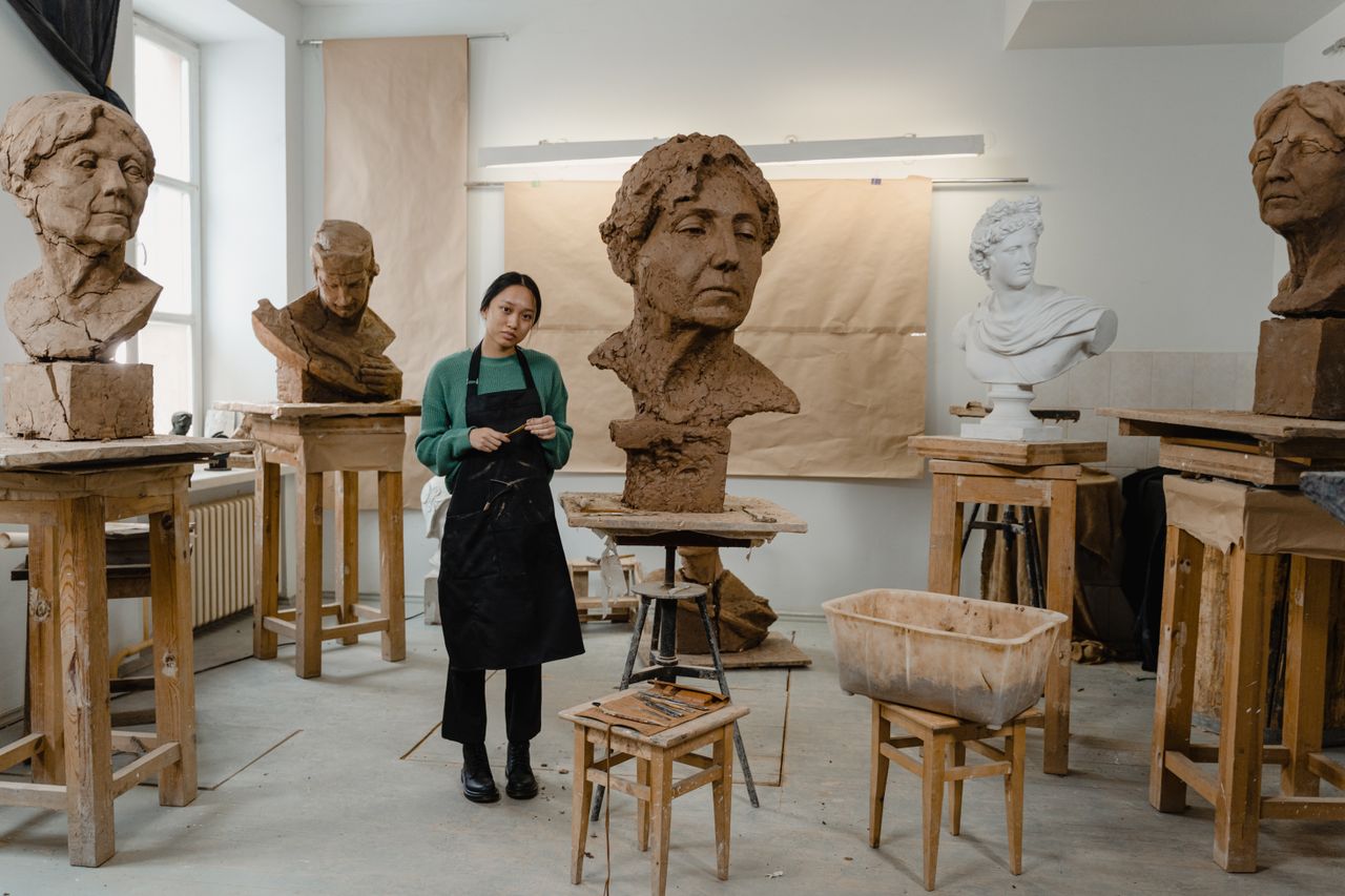 social-feed-img-04 Woman sculptor working on clay busts in a well-lit art studio, showcasing craftsmanship.
